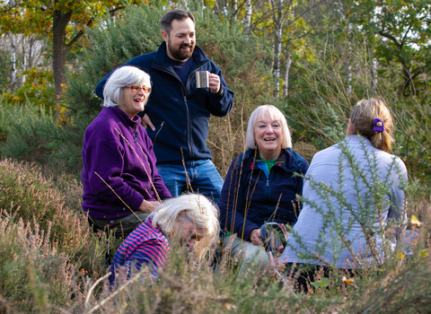 Group of people, aged 50+, smiling and holding cups in heath and wooded area