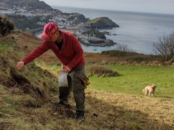 An individual spreading wildflower seed on a coastal hill, with a sea view behind.