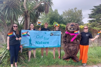 Five DWT staff and beaver costume standing with 'Out for Nature' banner and waving pride flags