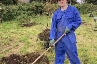 George, a man in a blue jumpsuit with a cap on, holding a garden tool and standing in a patch of grass