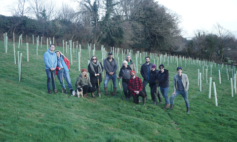 A group of people standing in a field with newly planting trees wrapped in protective plastic