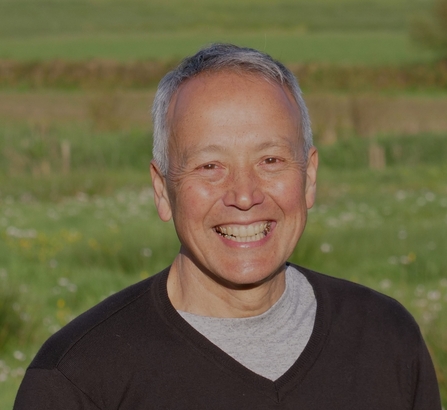 A man with short grey hair wearing a v-neck black jumper, standing in a field