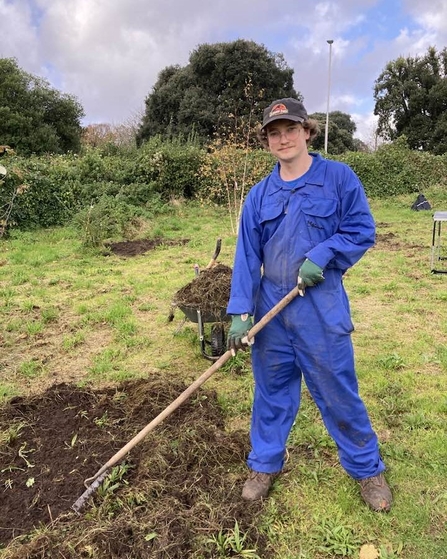 George, a man in a blue jumpsuit with a cap on, holding a garden tool and standing in a patch of grass