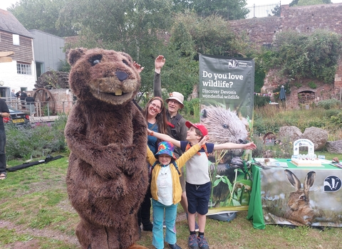 Family of two adults and two children stretching their arms next to large beaver costume in garden 